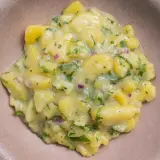 Ein cremiger Kartoffelsalat mit Speck, verfeinert mit frischer Petersilie und roten Zwiebeln, serviert in einer braunen Schale.