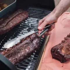 Spareribs werden mit Zangen auf dem Grill gewendet. Daneben liegen weitere Rippchen auf Butcher Paper.