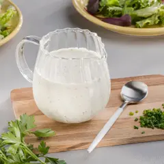 Ein Krug mit cremigem Ranch Dressing steht neben frischer Petersilie und Schnittlauch auf einem Holzbrett.