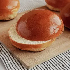 Ein aufgeschnittener, goldbrauner Brioche-Bun liegt auf einem Holzbrett. Weitere Buns sind im Hintergrund zu sehen.