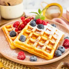 Zwei goldbraune Proteinwaffeln, bestäubt mit Puderzucker und garniert mit frischen Beeren und Johannisbeeren.