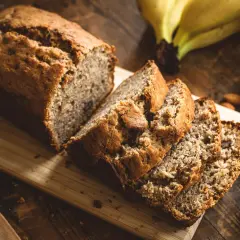 Ein saftiges Bananenbrot ohne Zucker, frisch aufgeschnitten auf einem Holzbrett. Daneben liegen reife Bananen und Mandeln.
