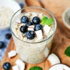 Ein Glas mit Overnight Oats Grundrezept, garniert mit Blaubeeren, Kokosraspeln und Minze. Daneben Chia-Samen und Haferflocken.