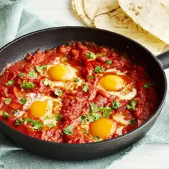 Herzhaftes Shakshuka Rezept in einer Gusseisenpfanne, mit vier pochierten Eiern in würziger Tomaten-Paprika-Sauce, garniert mit frischem Koriander.