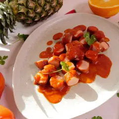 Ein Teller mit geschnittener Currywurst in Sauce, garniert mit Petersilie. Daneben Ananas, Orange, Erdbeeren und Tomaten – Zutaten für ein leckeres Currywurst Rezept.
