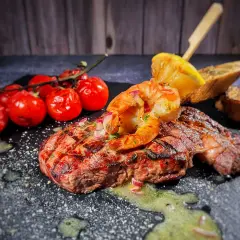 Perfektes Surf and Turf Rezept: saftiges Steak mit gegrillten Garnelen, Kirschtomaten, Zitrone und Brot auf dunklem Stein serviert.