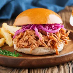 Ein saftiger Pulled Pork Burger mit Rotkohl und Pommes auf einem Holzplateau.
