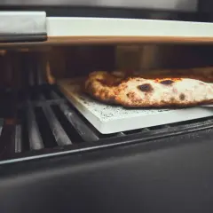 Eine köstliche Pizza wird auf einem Pizzastein im Gasgrill gebacken. Der Rand ist goldbraun und leicht angekohlt.