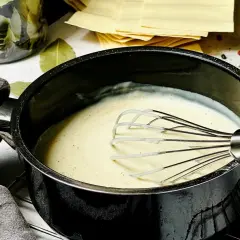 Cremige Béchamelsauce wird in einem Topf mit einem Schneebesen zubereitet. Daneben liegen Lasagneplatten und Gewürze.