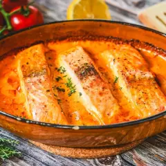 Saftige Lachsfilets in cremiger Lachs Sahne Sauce, garniert mit frischen Kräutern. Daneben Tomaten und eine Zitronenspalte.