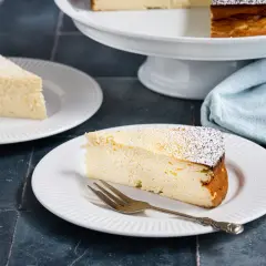 Ein saftiges Stück Quarkkuchen ohne Boden, bestäubt mit Puderzucker, auf einem Teller mit einer Gabel.