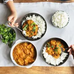 Drei Schalen mit Reis und leckeren indischen Gerichten, garniert mit Koriander. Eine Hand nimmt mit einem Löffel eine Portion Curry.