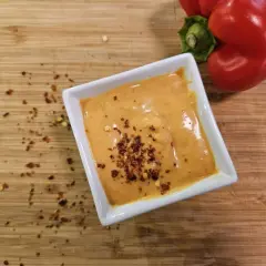 Cremige Chipotle-Sauce in einer weißen Schale, bestreut mit Chiliflocken. Daneben liegt eine rote Paprika auf einem Holzbrett.