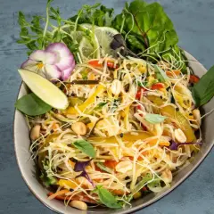Bunter Asia Salat mit Reisnudeln, Erdnüssen, Gemüse und frischen Kräutern, garniert mit Limettenspalten und einer essbaren Blüte.