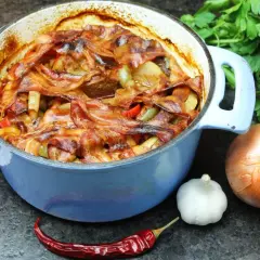 Herzhaftes Schichtfleisch im Backofen, bedeckt mit knusprigem Speck, daneben Zwiebel, Knoblauch und Chili.