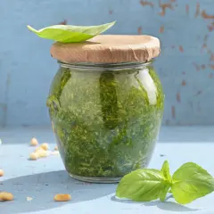 Frisches Pesto im Glas, garniert mit Basilikumblatt. Perfekt für dein nächstes Pesto Rezept. Pinienkerne und Parmesan liegen daneben.