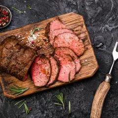 Saftiges Roastbeef, perfekt gegrillt und in Scheiben geschnitten, liegt auf einem Holzbrett. Gewürze und Rosmarin garnieren das Gericht.