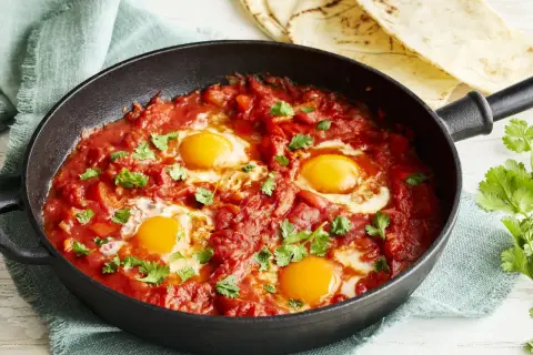 Herzhaftes Shakshuka Rezept in einer Gusseisenpfanne, mit vier pochierten Eiern in würziger Tomaten-Paprika-Sauce, garniert mit frischem Koriander.
