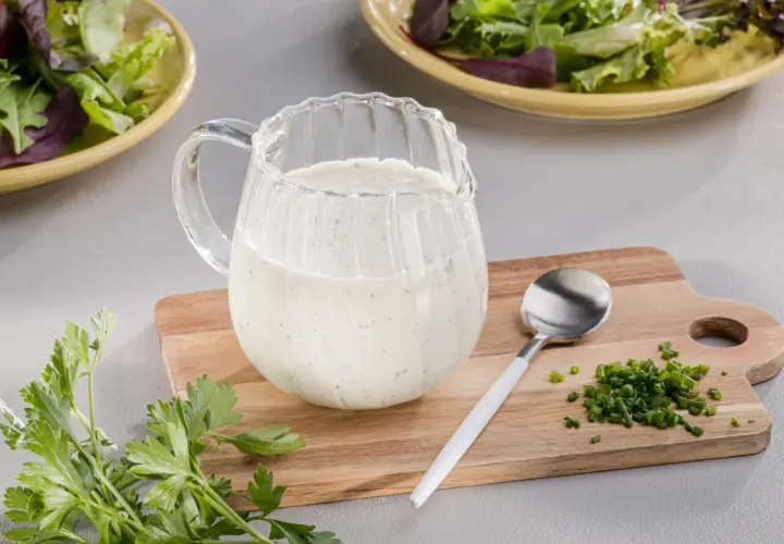 Ein Krug mit cremigem Ranch Dressing steht neben frischer Petersilie und Schnittlauch auf einem Holzbrett.