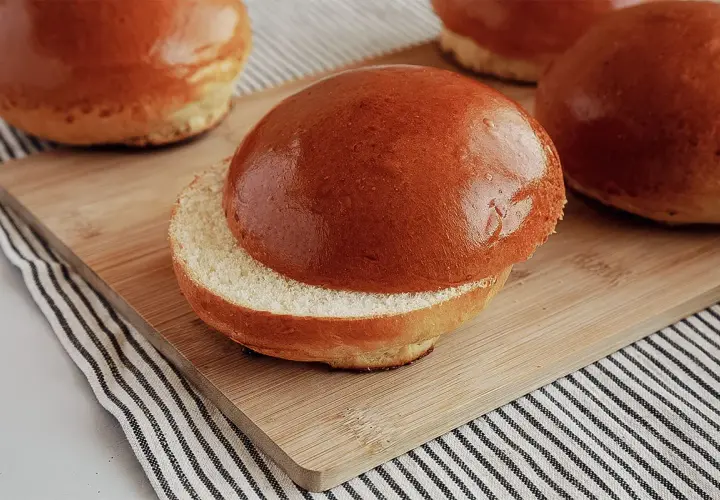 Ein aufgeschnittener, goldbrauner Brioche-Bun liegt auf einem Holzbrett. Weitere Buns sind im Hintergrund zu sehen.