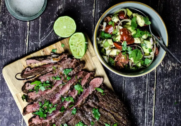 Perfekt gegrilltes Flanksteak, in Scheiben geschnitten, mit frischem Koriander und Limettenspalten, daneben ein bunter Salat.