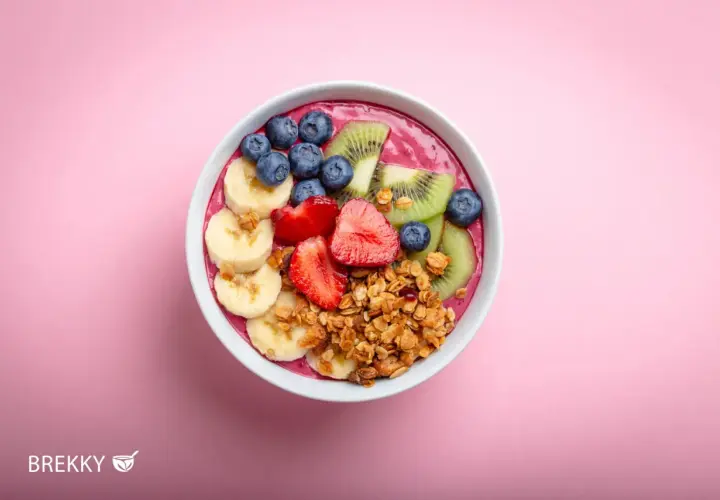 Ein farbenfrohes Acai Bowl Rezept mit Bananen, Erdbeeren, Blaubeeren, Kiwi und Granola auf rosa Hintergrund.