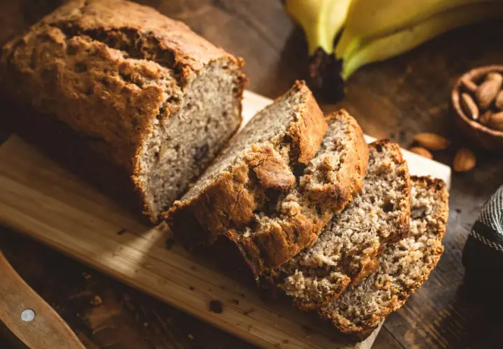 Ein saftiges Bananenbrot ohne Zucker, frisch aufgeschnitten auf einem Holzbrett. Daneben liegen reife Bananen und Mandeln.