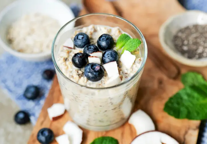 Ein Glas mit Overnight Oats Grundrezept, garniert mit Blaubeeren, Kokosraspeln und Minze. Daneben Chia-Samen und Haferflocken.