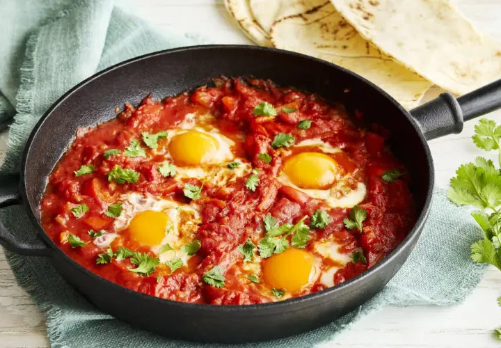 Herzhaftes Shakshuka Rezept in einer Gusseisenpfanne, mit vier pochierten Eiern in würziger Tomaten-Paprika-Sauce, garniert mit frischem Koriander.