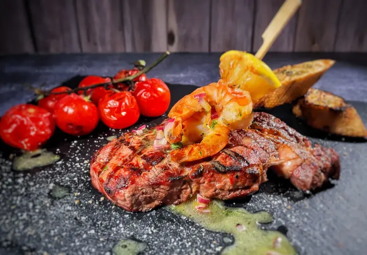 Perfektes Surf and Turf Rezept: saftiges Steak mit gegrillten Garnelen, Kirschtomaten, Zitrone und Brot auf dunklem Stein serviert.