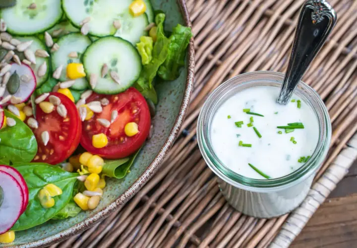 Frischer Salat mit Tomaten, Gurken und Radieschen neben einem Glas mit selbstgemachtem Joghurt-Dressing, garniert mit Schnittlauch.
