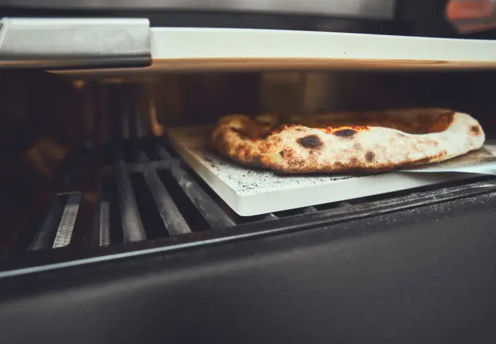 Eine köstliche Pizza wird auf einem Pizzastein im Gasgrill gebacken. Der Rand ist goldbraun und leicht angekohlt.