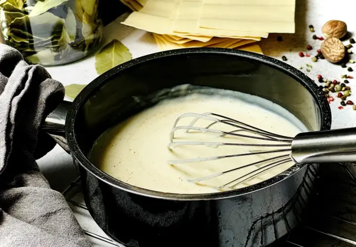 Cremige Béchamelsauce wird in einem Topf mit einem Schneebesen zubereitet. Daneben liegen Lasagneplatten und Gewürze.