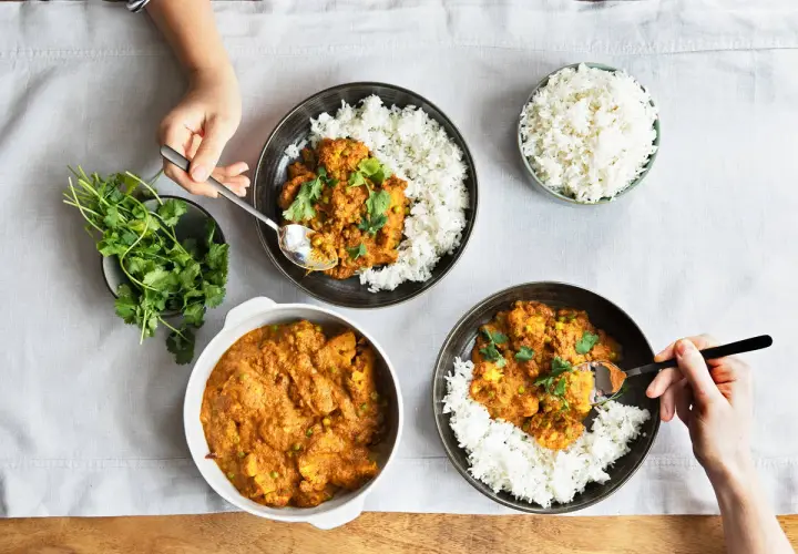 Drei Schalen mit Reis und leckeren indischen Gerichten, garniert mit Koriander. Eine Hand nimmt mit einem Löffel eine Portion Curry.