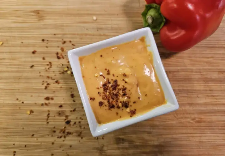 Cremige Chipotle-Sauce in einer weißen Schale, bestreut mit Chiliflocken. Daneben liegt eine rote Paprika auf einem Holzbrett.