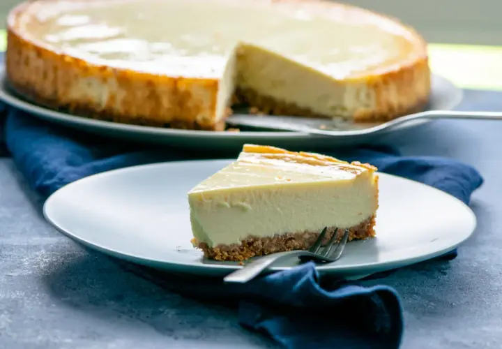 Ein Stück New York Cheesecake mit knusprigem Boden und cremiger Füllung auf einem Teller, daneben der ganze Kuchen.