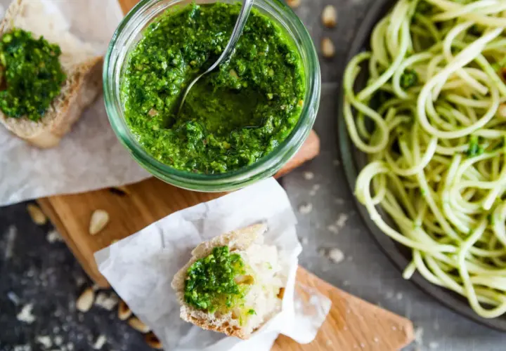 Frisches Basilikum-Pesto selber machen ist einfach! Hier ist ein Glas mit dem grünen Genuss, daneben Brot mit Pesto und eine Portion Spaghetti.