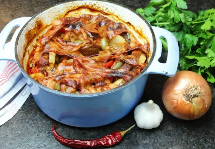 Herzhaftes Schichtfleisch im Backofen, bedeckt mit knusprigem Speck, daneben Zwiebel, Knoblauch und Chili.