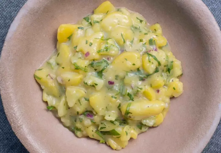 Ein cremiger Kartoffelsalat mit Speck, verfeinert mit frischer Petersilie und roten Zwiebeln, serviert in einer braunen Schale.