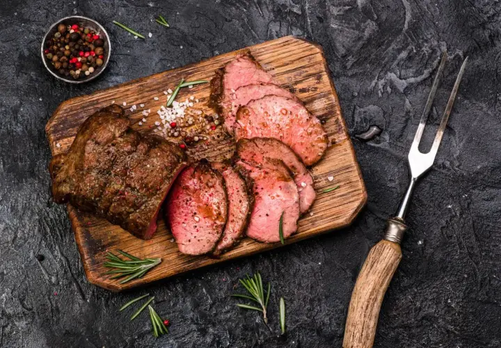 Saftiges Roastbeef, perfekt gegrillt und in Scheiben geschnitten, liegt auf einem Holzbrett. Gewürze und Rosmarin garnieren das Gericht.