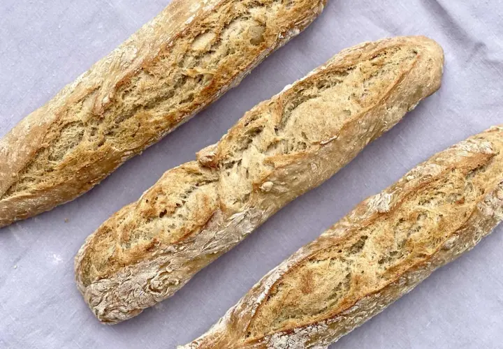 Drei frisch gebackene Baguettes mit knuspriger Kruste liegen auf einem grauen Tuch. Ein tolles Baguette Rezept zum Nachbacken!