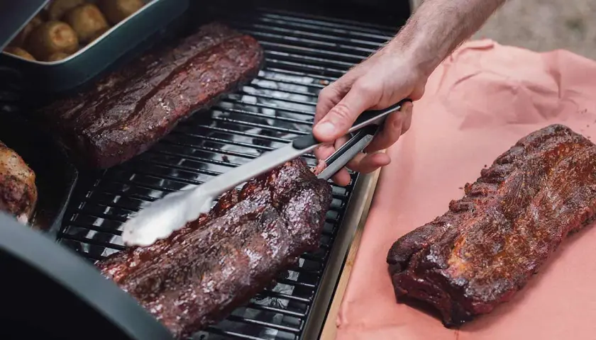 Spareribs werden mit Zangen auf dem Grill gewendet. Daneben liegen weitere Rippchen auf Butcher Paper.