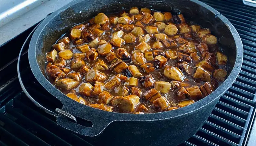 Leckere Currywurst-Stücke köcheln in einer würzigen Sauce in einem gusseisernen Dutch Oven auf dem Grill.