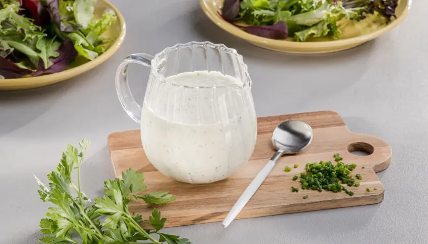 Ein Krug mit cremigem Ranch Dressing steht neben frischer Petersilie und Schnittlauch auf einem Holzbrett.