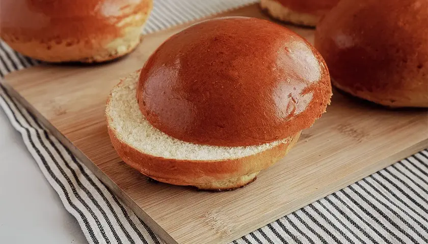 Ein aufgeschnittener, goldbrauner Brioche-Bun liegt auf einem Holzbrett. Weitere Buns sind im Hintergrund zu sehen.
