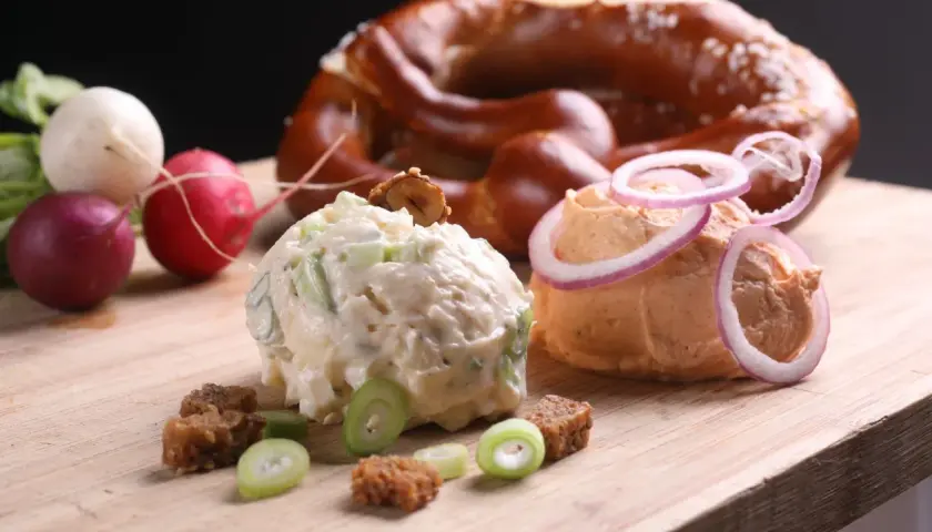 Zwei Portionen Obazda, garniert mit Zwiebelringen und Kräutern, daneben Radieschen und ein Brezel. Perfekt für ein Obazda Rezept!