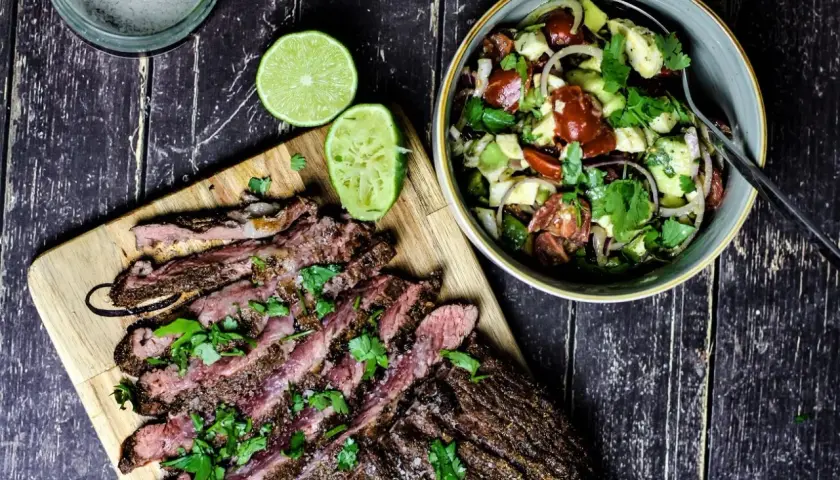 Perfekt gegrilltes Flanksteak, in Scheiben geschnitten, mit frischem Koriander und Limettenspalten, daneben ein bunter Salat.