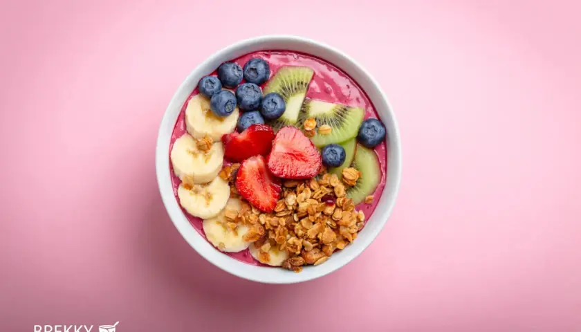 Ein farbenfrohes Acai Bowl Rezept mit Bananen, Erdbeeren, Blaubeeren, Kiwi und Granola auf rosa Hintergrund.