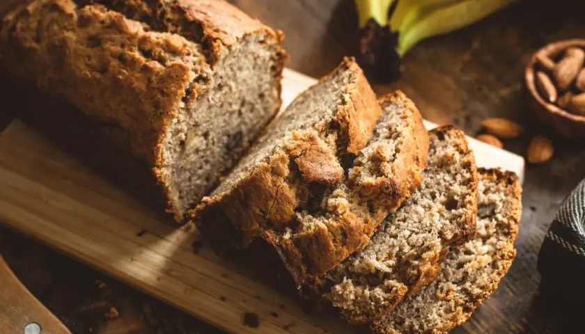 Ein saftiges Bananenbrot ohne Zucker, frisch aufgeschnitten auf einem Holzbrett. Daneben liegen reife Bananen und Mandeln.