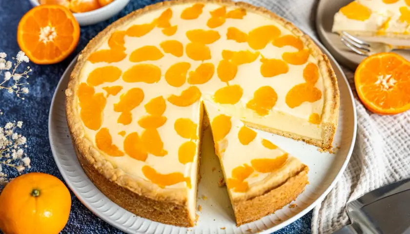 Ein saftiger faule weiber kuchen mit Mandarinenstücken, der auf einem Teller liegt. Daneben liegen frische Mandarinen und eine Tortenschaufel.